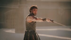 Paul Mescal as Lucius in Gladiator 2, with sword at the ready in fighting stance, clad in battle-worn Roman armor, with the arena blurred a bit in the back.