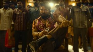 "Man in vibrant shirt and vest, seated on a wooden chair, surrounded by onlookers in a tense scene from a film.