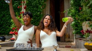 Two women in white dresses celebrating with a toast in a vibrant outdoor setting from the movie "Blink Twice."