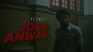 A man standing outside in a dimly lit scene with a large painted sign that reads 'Film Karya Joko Anwar.'(Grave Torture)