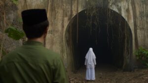 A man in the movie Grave Torture in a green shirt and a cap looks towards a small figure in white, standing at the entrance of a dark, overgrown tunnel.