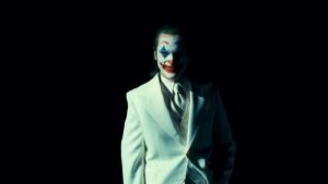 joaquin Phoenix as Joker in a white suit, standing against a dark background in a scene from 'Joker: Folie à Deux,' with his iconic makeup and a haunting expression.