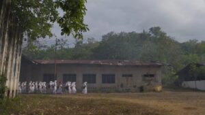 A group of figures in white robes walking towards an old, gray building in a foggy, open area "Grave Torture"