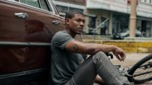 Scene from Rebel Ridge showing the main character sitting thoughtfully on the curb next to a vintage car, reflecting a moment of quiet in the movie.