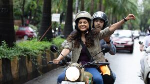 Scene from Sharmajee Ki Beti featuring a young woman and man enjoying a scooter ride through city streets.