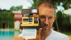  Man holding a vintage Polaroid camera by the poolside in a scene from the movie "Blink Twice."